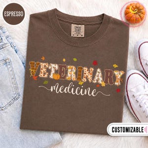 Puede incluir: Una camiseta color marrón espresso con el texto VETERINARY medicine en una fuente decorativa. Las letras están rellenas con patrones de temática otoñal que incluyen calabazas y hojas. La camiseta tiene cuello redondo y etiqueta Comfort Colors.