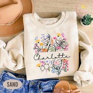 May include: Cream-colored sweatshirt featuring a floral and science-themed design. The design includes DNA strands, an atom symbol, and colorful flowers. The name "Charlotte" is written in cursive script below the graphic.