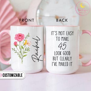 May include: A white ceramic mug with a pink handle and interior. The front features a floral design with pink and yellow flowers and the name "Rachel". The back of the mug says "IT'S NOT EASY TO MAKE 45 LOOK GOOD BUT CLEARLY I'VE NAILED IT".