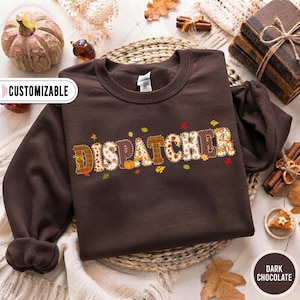 Op de afbeelding: Een donker chocoladebruin sweatshirt met ronde hals en de tekst DISPATCHER in een decoratief lettertype met herfstpatronen. De letters bevatten ruitjes, bloemmotieven en herfstbladeren in tinten oranje, geel en bruin.