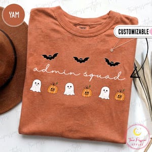 May include: A rust-colored t-shirt featuring a graphic print with three black bats, the white cursive text "admin squad", and a row of three white ghosts alternating with three orange jack-o'-lantern pumpkins across the center chest area.