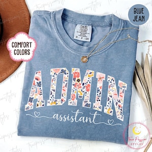 May include: Blue jean-colored t-shirt with the word "ADMIN" in floral print and the word "assistant" in script. The shirt has a crew neck and a Comfort Colors tag. A gold necklace with a pendant is draped on the shirt.