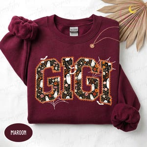 K&ouml;nnte beinhalten: Ein kastanienbraunes Sweatshirt mit Rundhalsausschnitt und dem Schriftzug GIGI in gro&szlig;en Blockbuchstaben. Die Buchstaben sind mit einem Muster aus wei&szlig;en Geistern, orangefarbenen und rosa Blumen sowie gr&uuml;nen Bl&auml;ttern auf schwarzem Grund gef&uuml;llt.