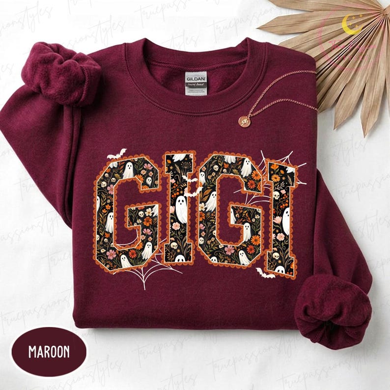 K&ouml;nnte beinhalten: Ein kastanienbraunes Sweatshirt mit Rundhalsausschnitt und dem Schriftzug GIGI in gro&szlig;en Blockbuchstaben. Die Buchstaben sind mit einem Muster aus wei&szlig;en Geistern, orangefarbenen und rosa Blumen sowie gr&uuml;nen Bl&auml;ttern auf schwarzem Grund gef&uuml;llt.