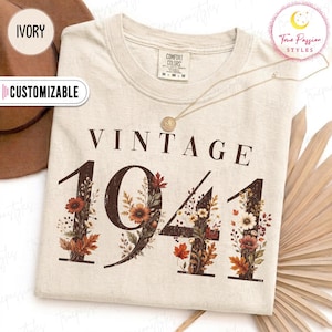 May include: An ivory short-sleeve t-shirt with a crew neckline. The graphic print features the text VINTAGE 1941 in dark brown, with the numbers filled with illustrations of autumn leaves, wildflowers, and berries in shades of orange and brown.
