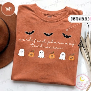 May include: A rust-colored crew neck t-shirt featuring the white text "certified pharmacy technician" centered on the chest. The design includes three black bats above the text and a row of three white ghosts alternating with three orange pumpkins below.