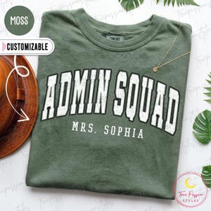 May include: A moss green crewneck t-shirt featuring the text ADMIN SQUAD in a large, distressed white collegiate font with a black outline, and the name MRS. SOPHIA printed in a smaller white sans-serif font directly underneath the main text.