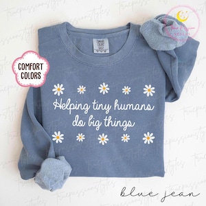 May include: A blue jean coloured long-sleeved shirt with the text "Helping tiny humans do big things" in white. The shirt features white daisies and the Comfort Colors brand tag. A blue scrunchie rests on the shirt.