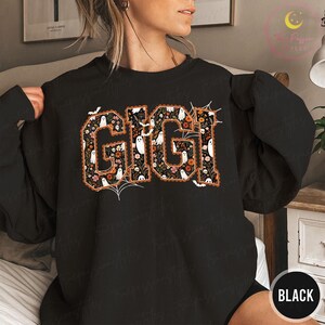 K&ouml;nnte beinhalten: Ein schwarzes Langarm-Sweatshirt mit dem Wort GIGI in gro&szlig;en Blockbuchstaben. Die Buchstaben sind mit einem Muster aus wei&szlig;en Geistern, orangefarbenen und rosa Blumen sowie kleinen Totenk&ouml;pfen auf schwarzem Grund gef&uuml;llt, akzentuiert durch Spinnweben.