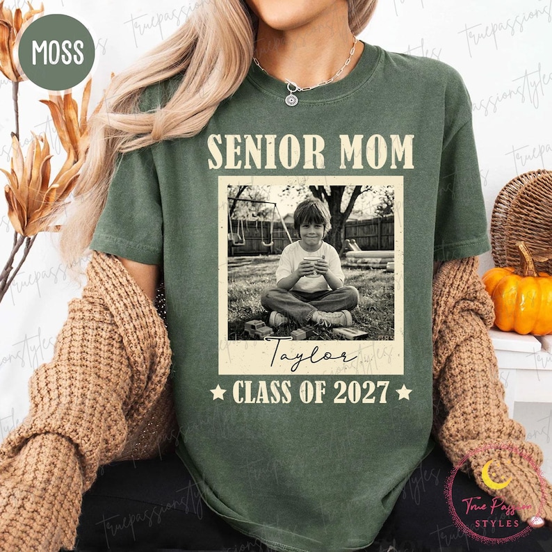 Może przedstawiać: Zielona koszulka w kolorze mchu z napisem "SENIOR MOM" nad czarno-białym zdjęciem dziecka. Zdjęcie jest podpisane "Taylor" i "CLASS OF 2027" z gwiazdkami. Koszulka ma swobodny styl.