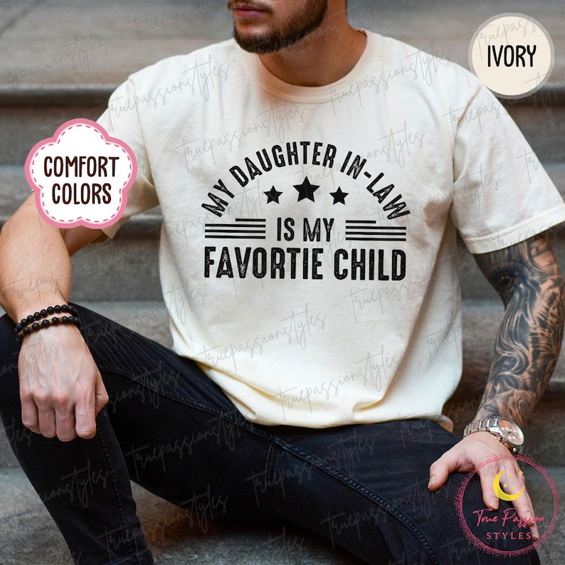 May include: Cream-coloured t-shirt featuring the text "MY DAUGHTER IN-LAW IS MY FAVORITE CHILD" in black. The shirt has a round neck and short sleeves. The words "COMFORT COLORS" are in a pink circle on the left side of the shirt.