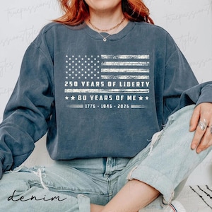 May include: A denim blue sweatshirt with a distressed American flag design. The flag graphic includes the text "250 YEARS OF LIBERTY" and "80 YEARS OF ME" with the years 1776-1946-2026. The word "denim" is printed at the bottom.