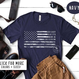 May include: A navy blue t-shirt with a distressed white American flag design. The text "BRISKET BUTTS RIBS & FREEDOM" is printed on the flag.