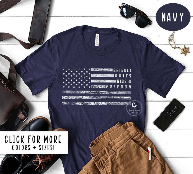 May include: A navy blue t-shirt with a distressed white American flag design. The text "BRISKET BUTTS RIBS & FREEDOM" is printed on the flag.