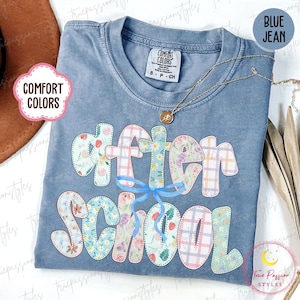 Könnte beinhalten: Ein blaues Comfort Colors T-Shirt mit der Aufschrift "After School" in einem floralen und karierten Design. Eine goldene Halskette mit Anhänger ist auf dem Shirt drapiert. Das Shirt ist gefaltet und auf einer Oberfläche ausgestellt.