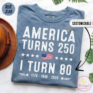 May include: Blue jean-colored t-shirt with the text "AMERICA TURNS 250" and "I TURN 80" in white. The shirt includes an American flag graphic and the years 1776-1946-2026. The shirt is customizable.