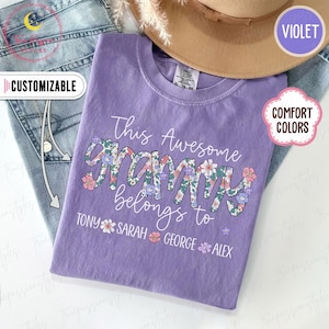 May include: A lilac-colored t-shirt with the text "This Awesome Grammy belongs to" and names below. The word "Grammy" is in a floral pattern. The shirt is next to a pair of jeans and a tan hat.