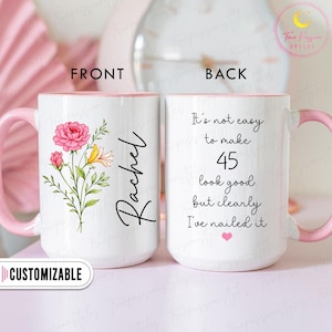 May include: White ceramic mug with a pink handle and interior. The front features a floral design with pink and yellow flowers and the name "Rachel". The back of the mug has the text "It's not easy to make 45 look good but clearly I've nailed it."
