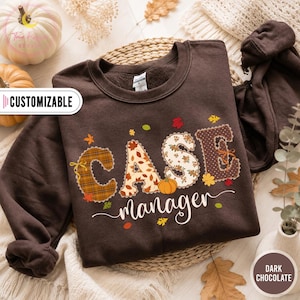 Op de afbeelding: Een donkerchocoladebruine sweater met ronde hals en de tekst CASE MANAGER in grote, gedessineerde letters. De letters zijn versierd met ruitpatronen, herfstbladeren en bloemmotieven, geaccentueerd door een kleine oranje pompoen en blaadjes.
