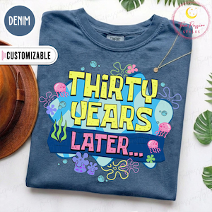 Op de afbeelding: Een denimblauw t-shirt met een grafische print met de gele tekst "THIRTY YEARS" boven de roze tekst "LATER..." op een blauwe achtergrond. Het ontwerp bevat illustraties van roze kwallen, groen zeewier en diverse kleurrijke zeebloemen.