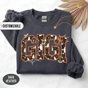 K&ouml;nnte beinhalten: Ein dunkelgrau meliertes Sweatshirt mit Rundhalsausschnitt und dem Schriftzug GIGI in gro&szlig;en Blockbuchstaben. Die Buchstaben sind mit einem Muster aus wei&szlig;en Geistern, orangefarbenen und rosa Blumen sowie Totenk&ouml;pfen auf schwarzem Grund gef&uuml;llt.
