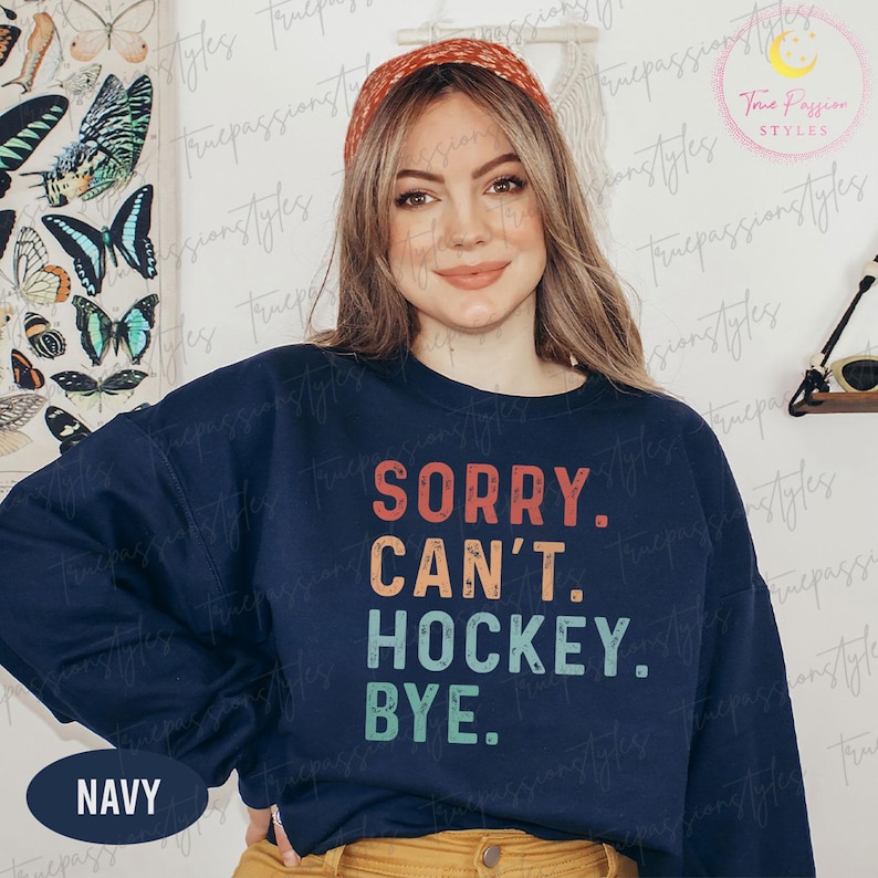 May include: Navy blue sweatshirt with the text "SORRY. CAN'T. HOCKEY. BYE." in orange, teal, and blue. The sweatshirt has long sleeves and a crew neck. A navy blue oval with the word "NAVY" is at the bottom left.