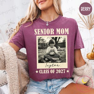 Może przedstawiać: Koszulka w kolorze jagodowym z napisem "SENIOR MOM" nad czarno-białym zdjęciem dziecka. Zdjęcie jest podpisane "Taylor" i "CLASS OF 2027". Koszulka ma okrągły dekolt.