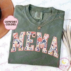 Può includere: T-shirt verde oliva con la scritta "MEMA" in grandi lettere a fantasia floreale bordate di corallo. La maglietta ha uno scollo tondo e un'etichetta "Comfort Colors". Una delicata collana con un ciondolo è appoggiata sulla maglietta.