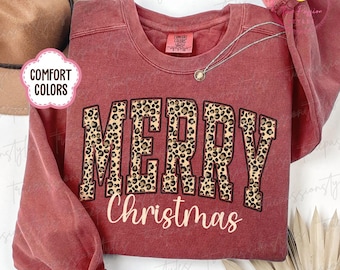 May include: Crimson red sweatshirt with the words "MERRY Christmas" in a leopard print design. The sweatshirt has long sleeves and a crew neck. A silver necklace with a small pendant is draped on the sweatshirt.