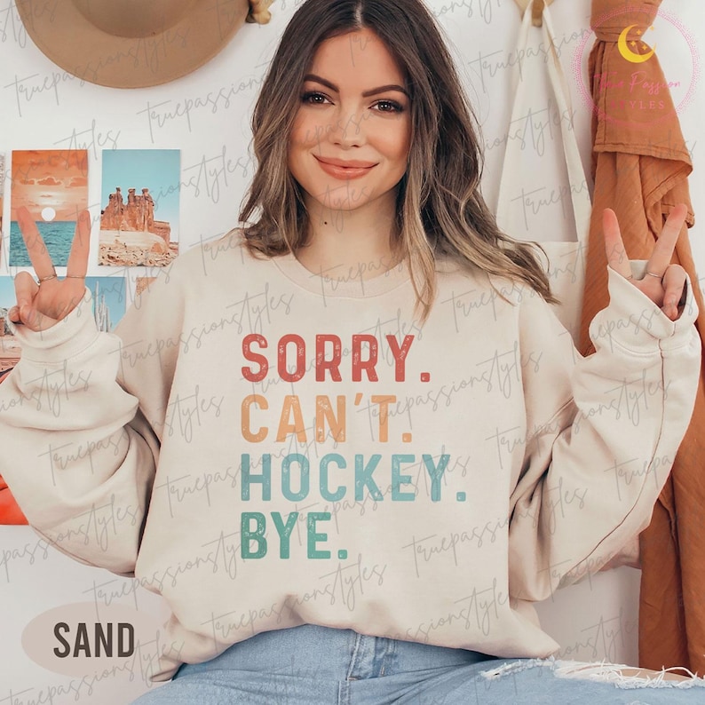 May include: A sand-coloured sweatshirt with the text "SORRY. CAN'T. HOCKEY. BYE." in red, orange, teal, and green. The word "SAND" is printed at the bottom. The sweatshirt is a casual style.