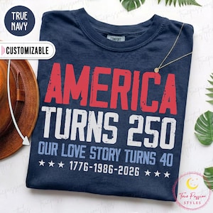 Könnte beinhalten: Ein marineblaues T-Shirt mit abgenutztem rotem und weißem Text, der AMERICA TURNS 250 lautet, gefolgt von OUR LOVE STORY TURNS 40 und den Jahren 1776-1986-2026 mit kleinen weißen Sternen in der untersten Zeile des Grafikdesigns.