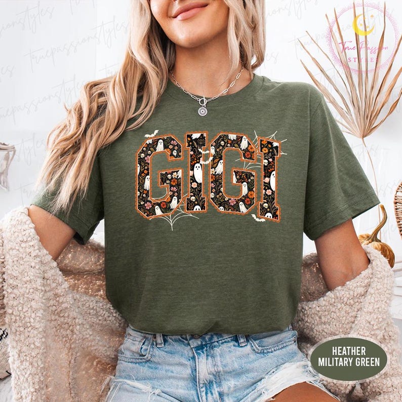K&ouml;nnte beinhalten: Ein kurz&auml;rmeliges T-Shirt in heather military green mit dem Schriftzug GIGI in gro&szlig;en Blockbuchstaben. Die Buchstaben sind mit einem Muster aus kleinen wei&szlig;en Geistern, orangefarbenen Blumen und schwarzem Laub gef&uuml;llt, erg&auml;nzt durch Spinnweben.