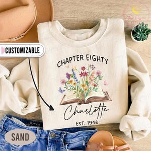 May include: Sand-colored sweatshirt with the text "Chapter Eighty" above an open book illustration with colorful flowers and butterflies. Below the book is the name "Charlotte" and "EST. 1946". The word "Customizable" is on the left.