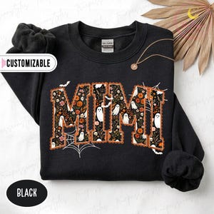 Könnte beinhalten: Ein schwarzes Sweatshirt mit Rundhalsausschnitt und dem Wort MIMI in großen Blockbuchstaben. Die Buchstaben sind mit einem Muster aus Geistern, orangefarbenen Blumen und Laub gefüllt. Das Design enthält weiße Spinnennetze und Fledermäuse.