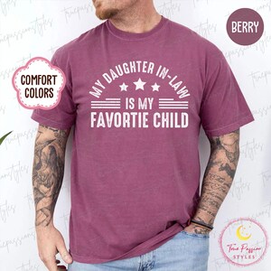 May include: A berry-coloured t-shirt with the text "MY DAUGHTER IN-LAW IS MY FAVORITE CHILD" in white. The shirt has a crew neck and short sleeves. The words "COMFORT COLORS" are in a pink cloud-shaped bubble.