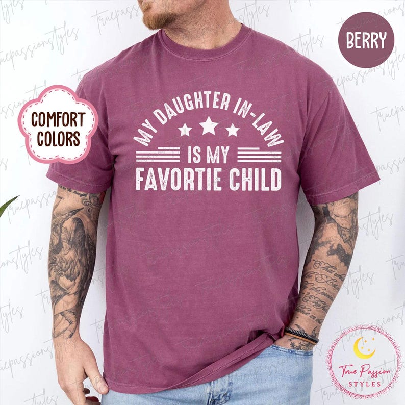 May include: A berry-coloured t-shirt with the text "MY DAUGHTER IN-LAW IS MY FAVORITE CHILD" in white. The shirt has a crew neck and short sleeves. The words "COMFORT COLORS" are in a pink cloud-shaped bubble.