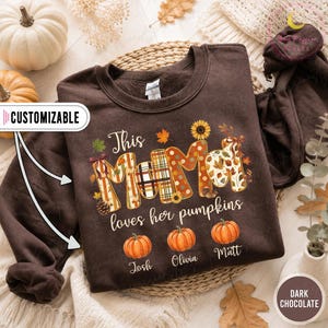 Könnte beinhalten: Ein dunkelbraunes Sweatshirt mit Rundhalsausschnitt und dem weißen Schriftzug "This MAMA loves her pumpkins". Das Wort "MAMA" ist mit herbstlichen Mustern gefüllt. Darunter befinden sich drei orangefarbene Kürbisillustrationen mit den Namen Josh, Olivia und Matt.