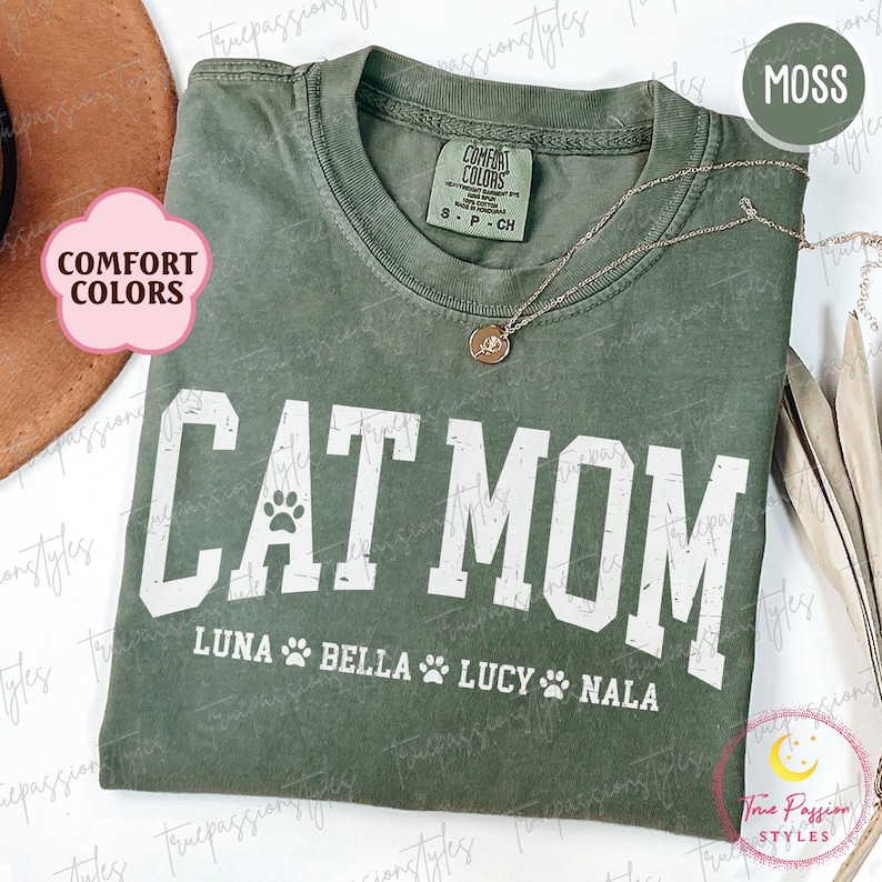 May include: Moss green Comfort Colors t-shirt with the text "CAT MOM" in white block letters. Below the text are the names Luna, Bella, Lucy, and Nala. A silver necklace with a pendant is draped on the shirt.