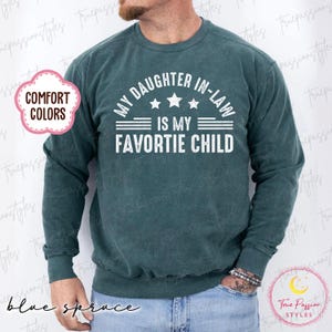May include: Blue spruce-coloured sweatshirt with the text "MY DAUGHTER IN-LAW IS MY FAVORITE CHILD" in white. The sweatshirt has long sleeves and a crew neck. The words "COMFORT COLORS" are in a pink cloud-shaped graphic.