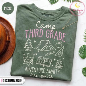May include: A moss green crew neck t-shirt featuring the white text Camp, pink text THIRD GRADE, and white text ADVENTURE AWAITS Mrs. Smith. The graphic includes a pine tree, tent, apple, crayon, ruler, folding chair, open book, and campfire.