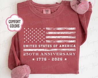 Puede incluir: Sudadera de color carmesí con un diseño de bandera estadounidense desgastada. El gráfico de la bandera incluye el texto "UNITED STATES OF AMERICA" y "250TH ANNIVERSARY 1776-2026". Una scrunchie está colocada en el hombro.