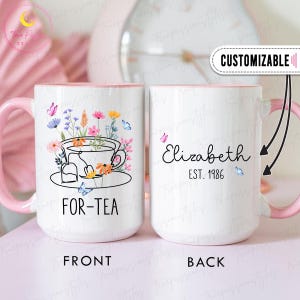 Peut inclure: Mug en céramique blanche avec une anse et un bord roses. Le devant présente un motif de tasse à thé floral avec le texte "FOR-TEA". Le dos de la tasse porte le nom "Elizabeth" et "EST. 1986". Des papillons et des fleurs décorent la tasse.