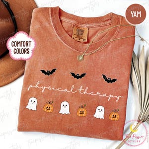 May include: A yam-colored Comfort Colors t-shirt with a crew neck and short sleeves. The shirt features a gold necklace and a design of black bats, white ghosts, and orange pumpkins. The words "physical therapy" are written in a script font.