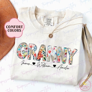 May include: Cream-colored t-shirt with the word "GRANNY" in a floral and leopard print design. Below the word are the names "James", "William", and "Amelia". A gold necklace is draped on the shirt. The shirt has a "Comfort Colors" tag.