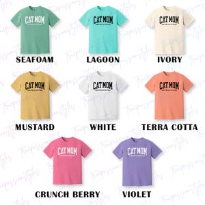 May include: A selection of short-sleeved t-shirts in a range of colours, each displaying the text "CAT MOM" in a curved, bold font. The shirts are available in seafoam, lagoon, ivory, mustard, white, terra cotta, crunch berry, and violet.