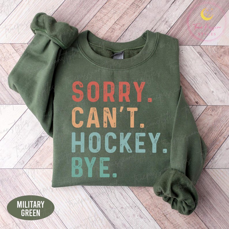 May include: A military green sweatshirt featuring the text "SORRY. CAN'T. HOCKEY. BYE." in red, orange, teal, and blue. The sweatshirt has a crew neck and long sleeves. The words "MILITARY GREEN" are at the bottom.