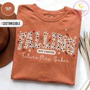 May include: A rust-colored crew neck t-shirt featuring the word FALLING in large block letters filled with a floral pattern of autumn leaves and flowers. Below this, the text reads INTO FOREVER and Future Mrs. Baker in white script.
