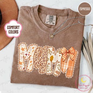 Könnte beinhalten: Espressofarbenes Comfort Colors T-Shirt mit dem Wort "MAMA" in dekorativer Schrift. Die Buchstaben sind mit floralen, Leopardenmuster und Karomustern verziert. Eine silberne Halskette mit Anhänger ist sichtbar.