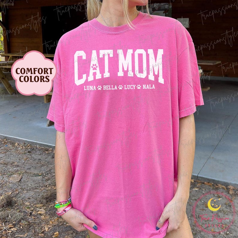 May include: A bright pink t-shirt featuring the text "CAT MOM" in white, bold lettering. Underneath, the names "LUNA", "BELLA", "LUCY", and "NALA" are displayed, each with a paw print. The shirt is a comfort colors style.