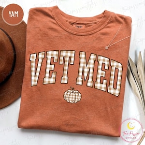 Può includere: Una t-shirt a maniche corte color ruggine con la scritta VET MED in un motivo a quadretti vichy beige e bianco. Sotto il testo si trova una piccola grafica a forma di zucca con lo stesso motivo. La maglietta ha un girocollo e un'etichetta.